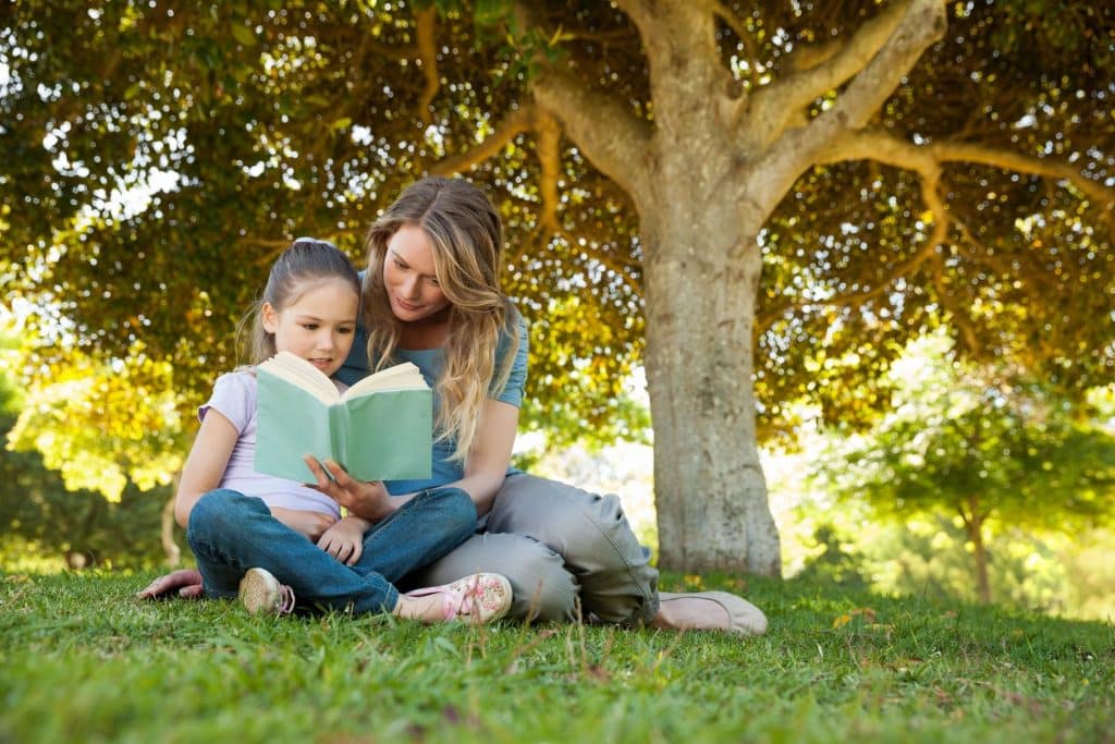 Mother and daughter reading a book together outdoors under a tree